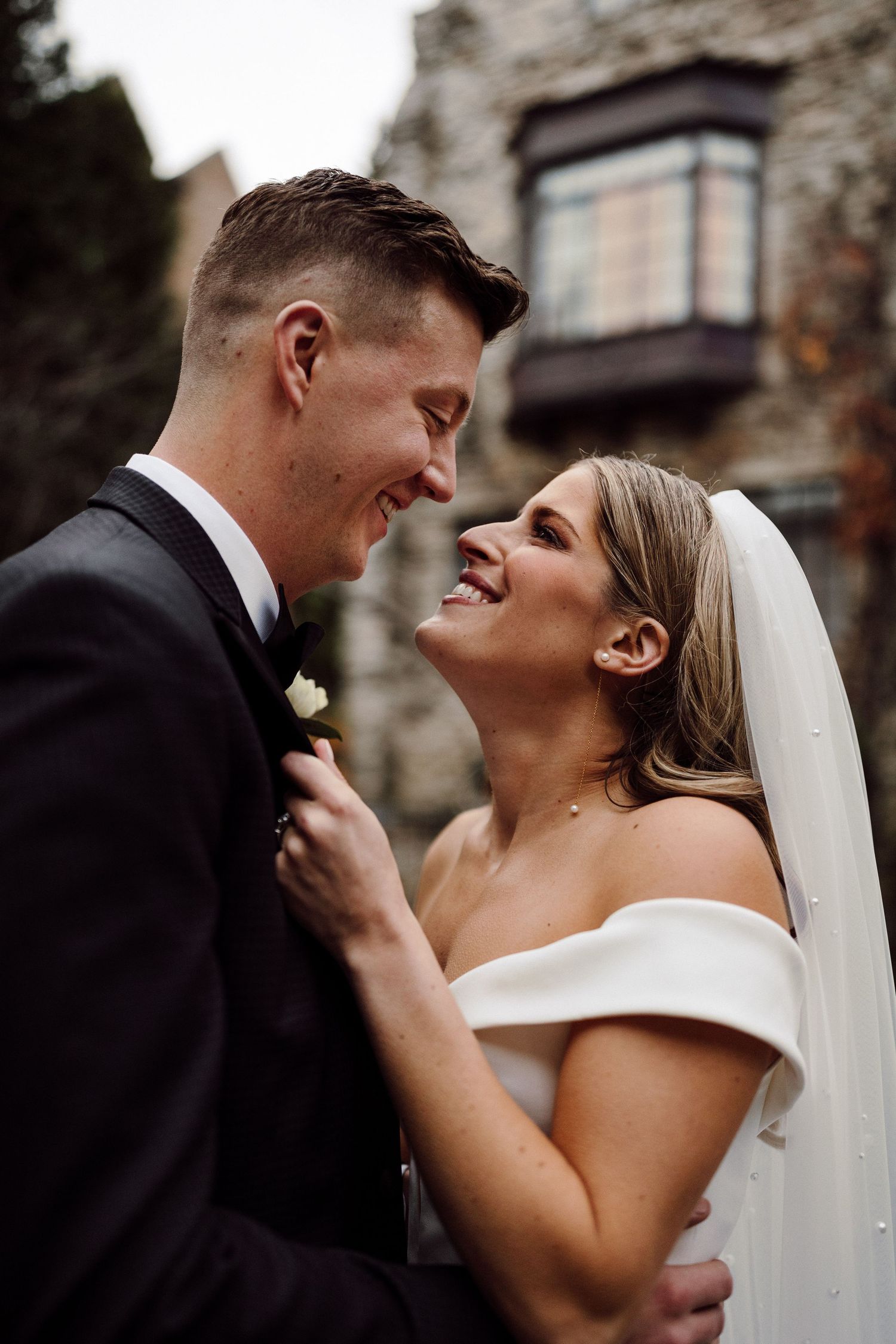 Smiling bride in off-shoulder gown and veil gazes at groom in black tuxedo outside stone wedding venue.