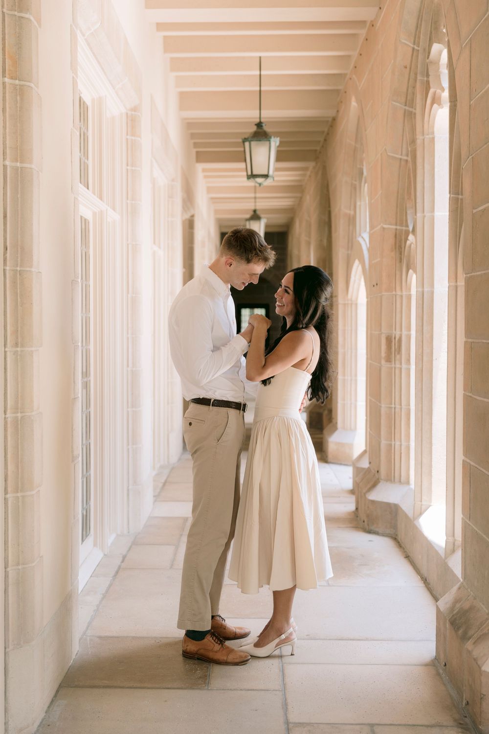 Couple smiling and holding hands in a elegant stone colonnade, woman in cream midi dress laughing joyfully.