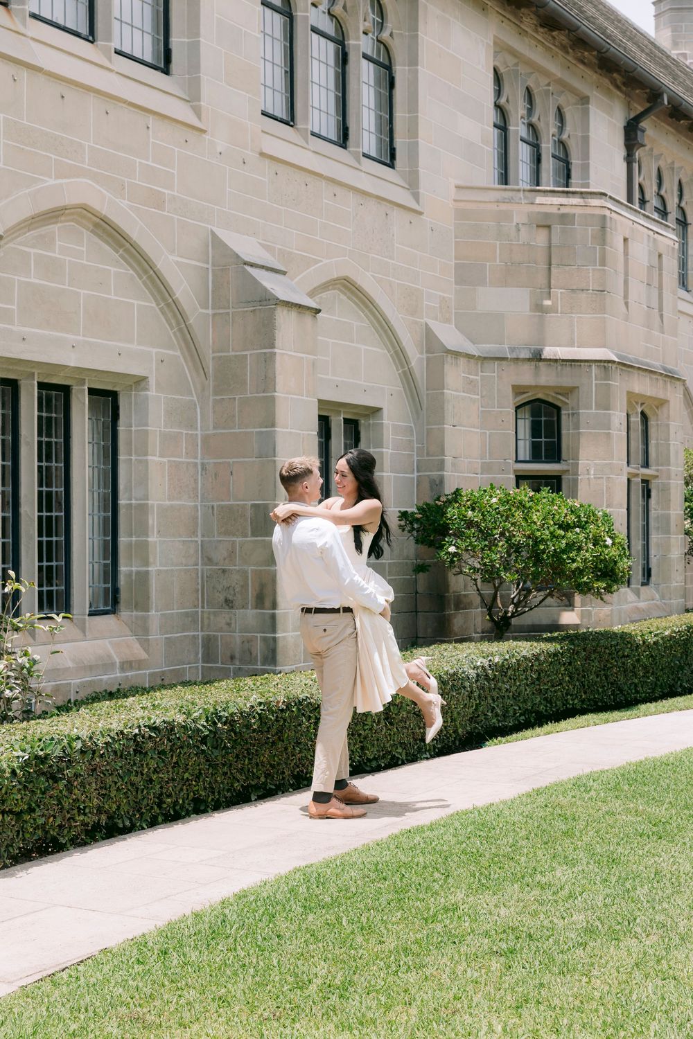 Couple embracing outside a historic stone building, man lifting woman on a sunny day near manicured hedges.