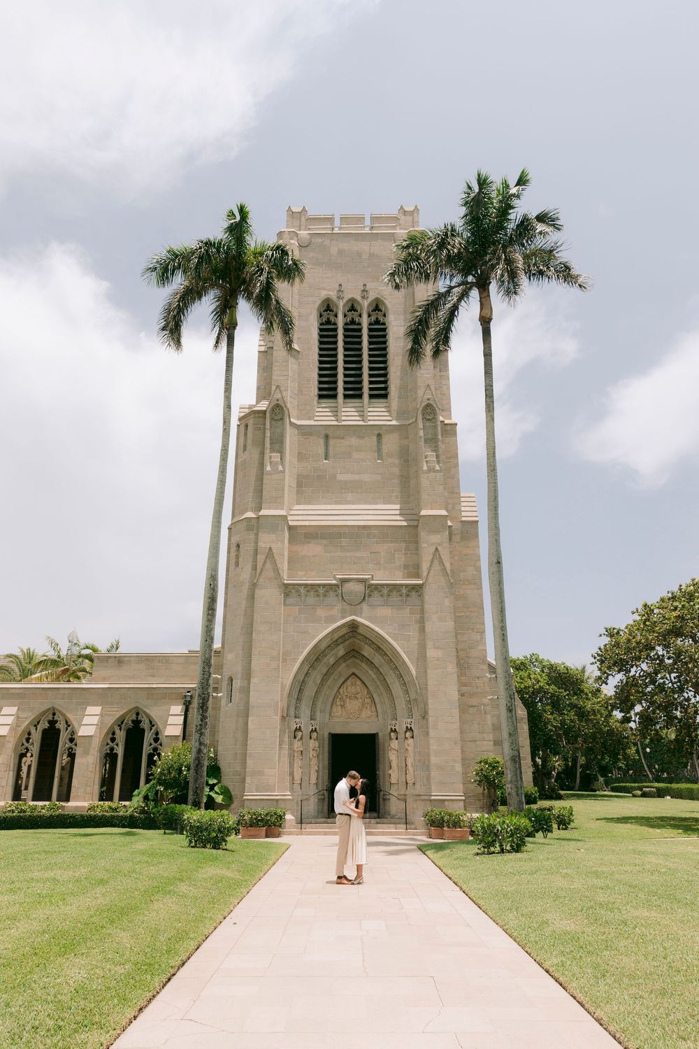 Couple embracing at entrance of historic Gothic church tower with palm trees and manicured lawn.