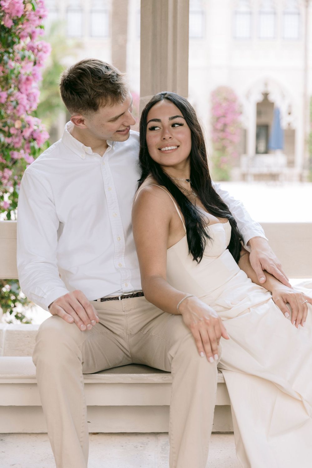 A couple sits together on a white bench surrounded by pink flowers, the woman in a cream dress smiling warmly at the camera.