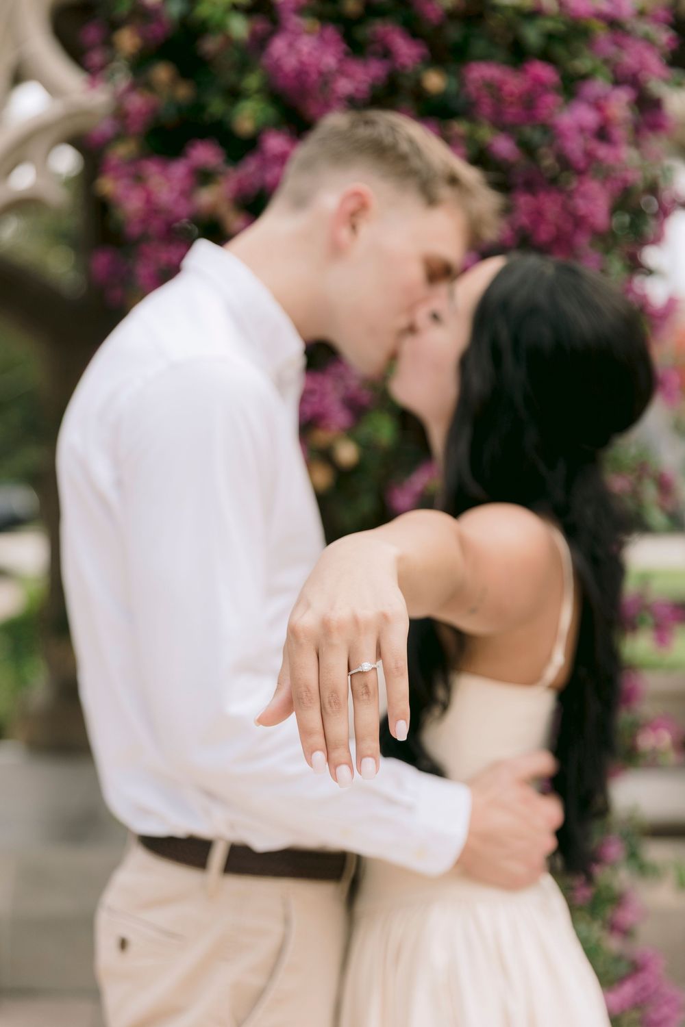 Engaged couple kissing in front of purple flowers, woman displaying diamond engagement ring on her hand.