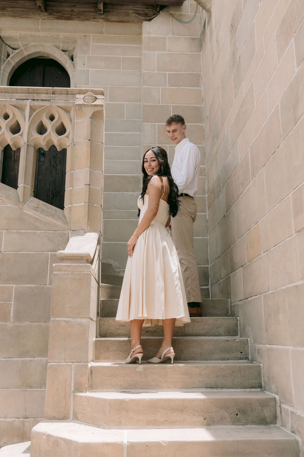 Couple posing on stone steps of historic building, woman in elegant cream midi dress with heels, man in white shirt behind her.