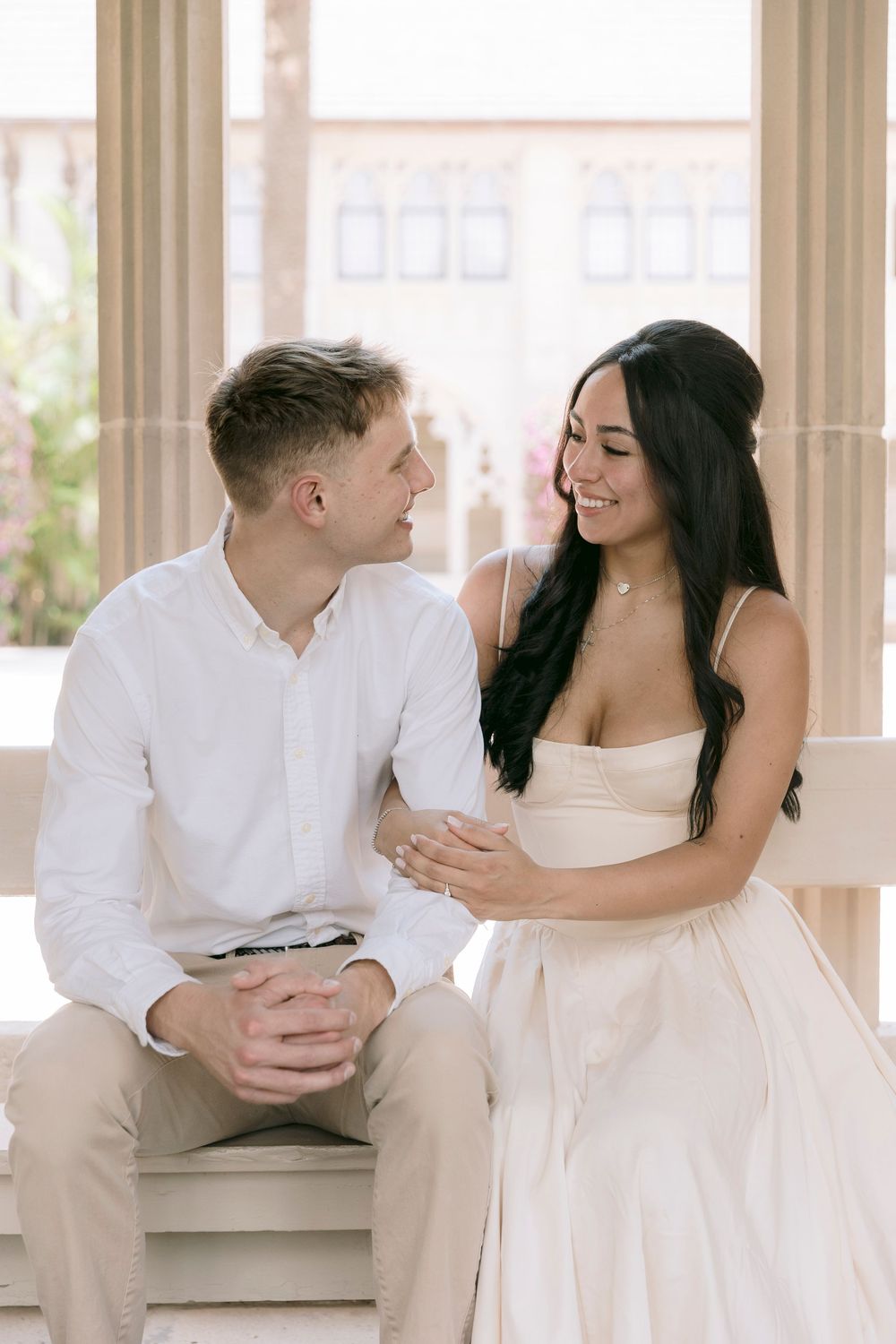 A couple in elegant white attire sits together on a bench, smiling at each other in a bright, classic setting.