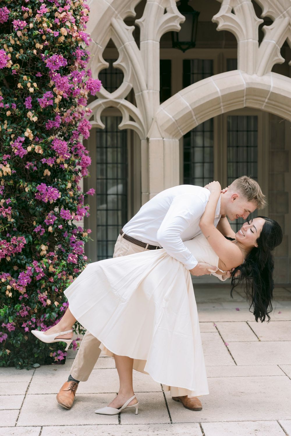 Couple sharing a romantic dip kiss in front of Gothic architecture with blooming flowers.
