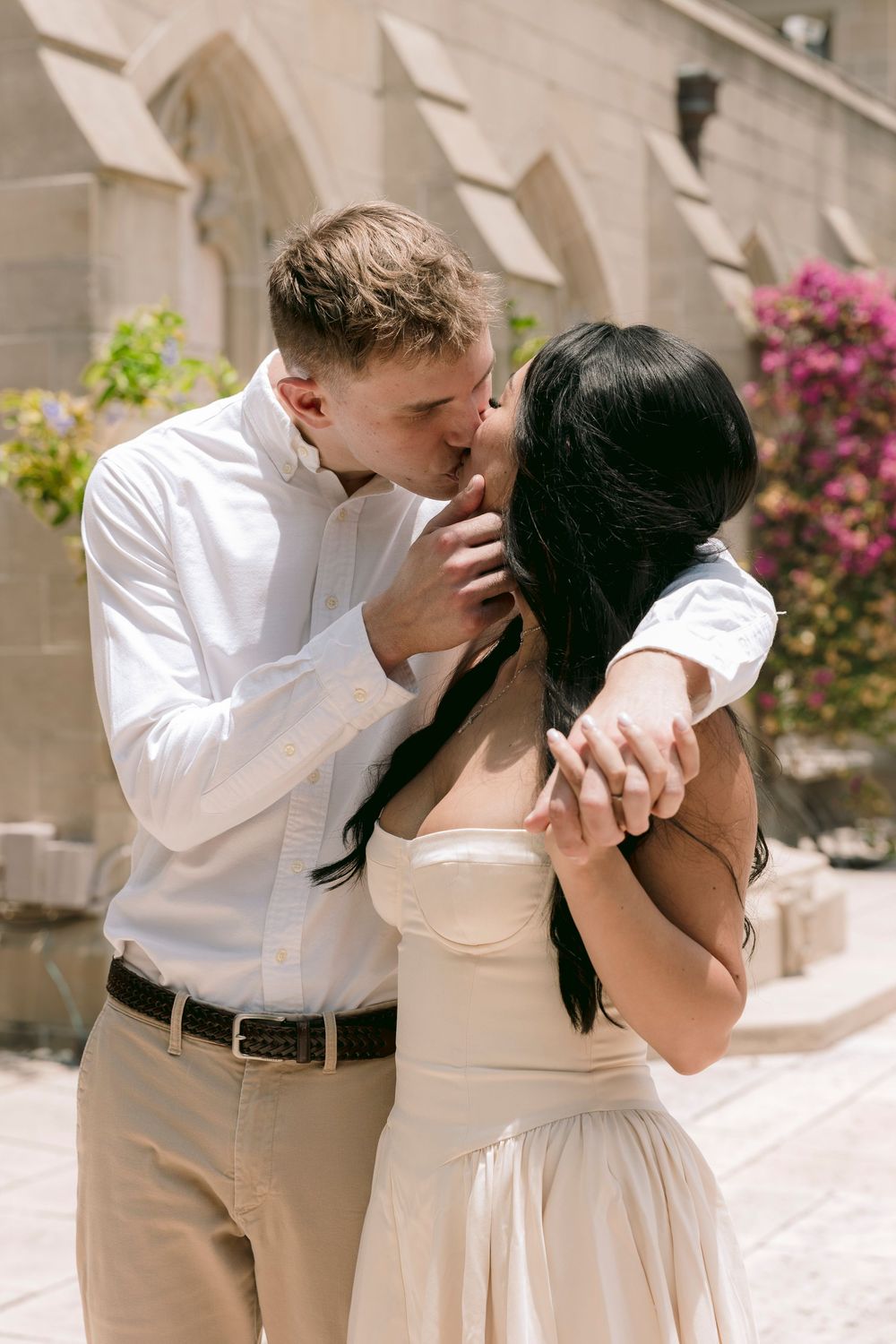 Couple sharing a romantic kiss outdoors near a stone building, man in white shirt and woman in elegant cream dress.