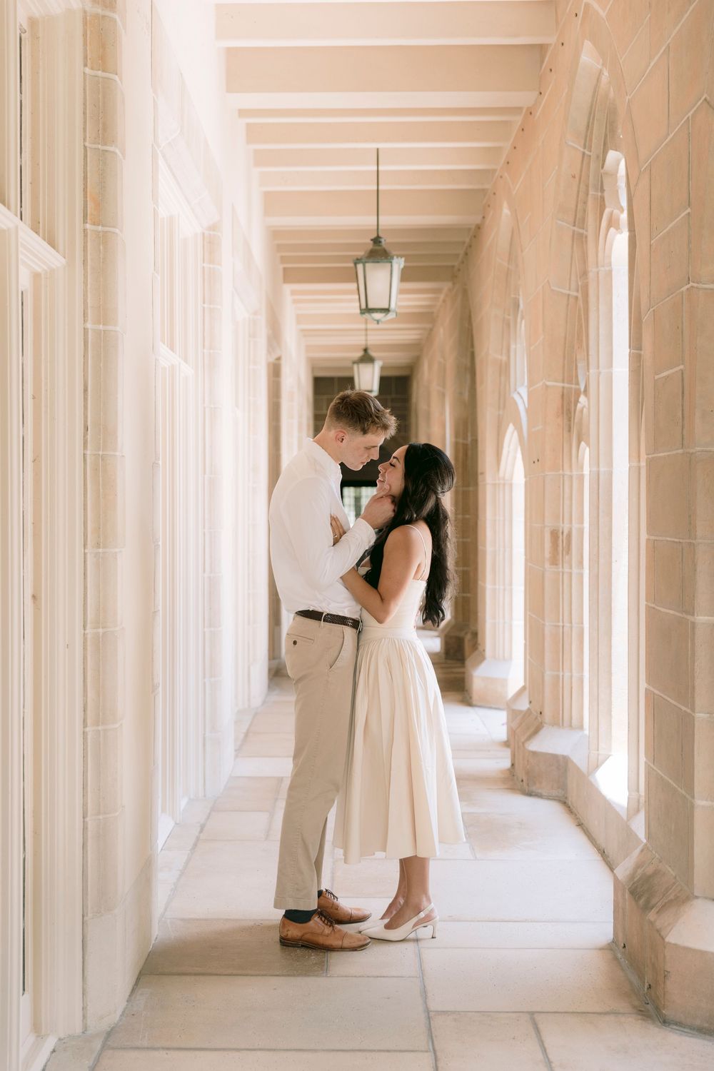 Couple embracing tenderly in a sunlit stone archway corridor, woman in cream midi dress and man in white shirt.