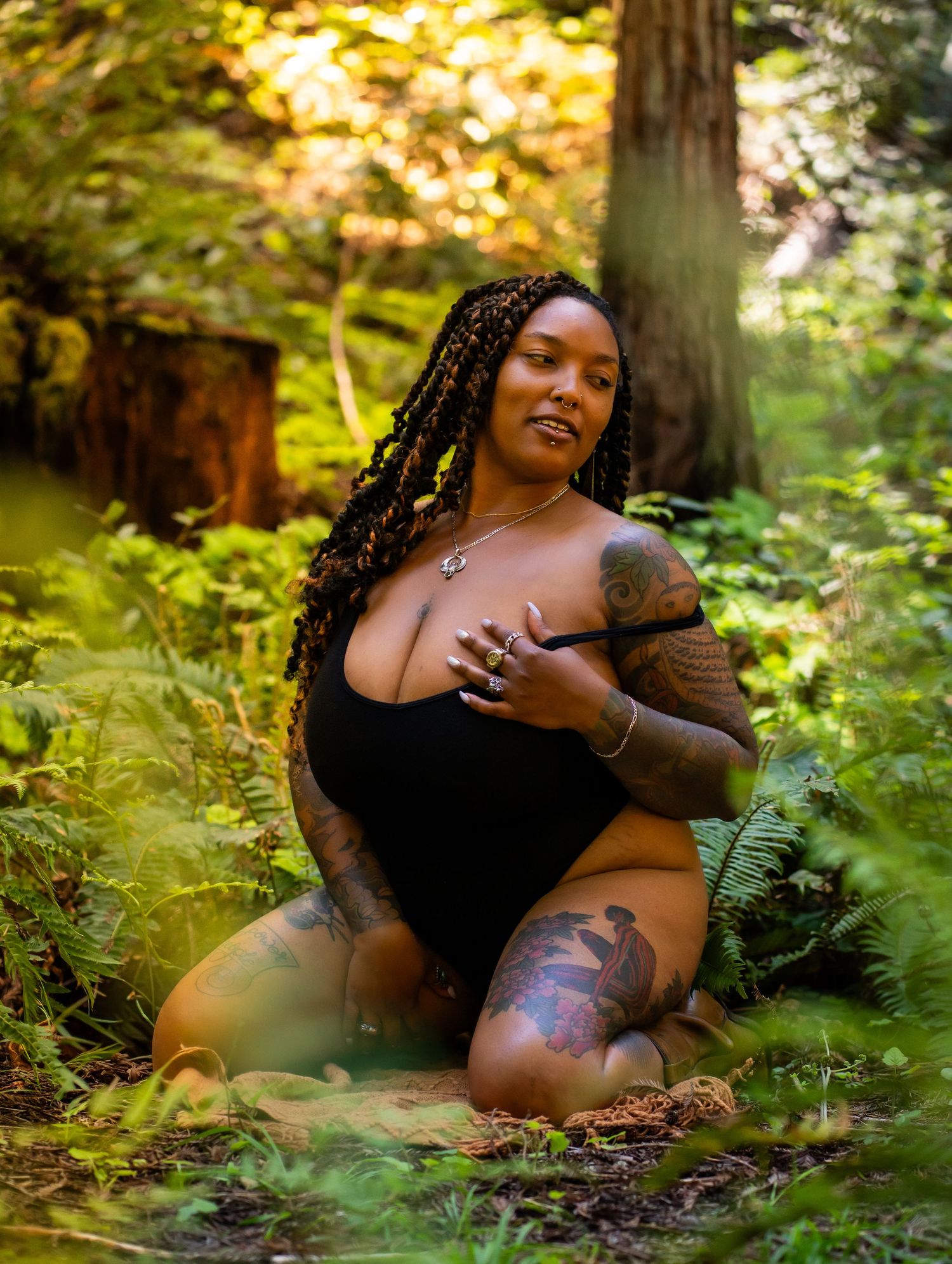 A tattooed woman with braids poses in a black bodysuit among lush green forest ferns and trees.