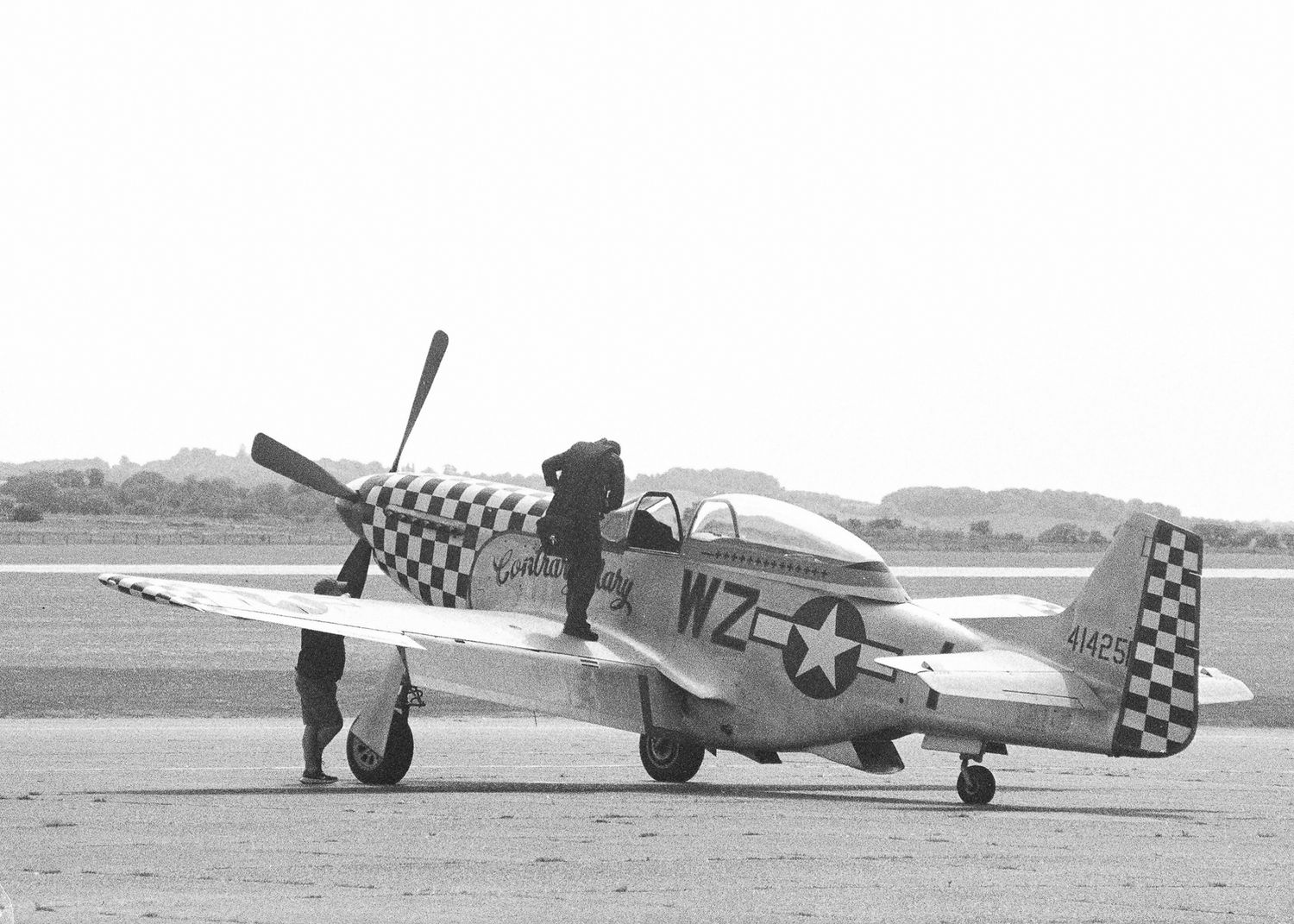 Vintage WWII P-51 Mustang fighter plane with checkered nose art and WZ markings on a runway in black and white photo.