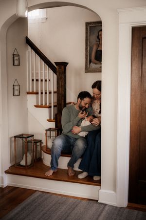 A couple sits on staircase steps in a cozy home, lovingly cradling their newborn baby beneath an arched doorway.