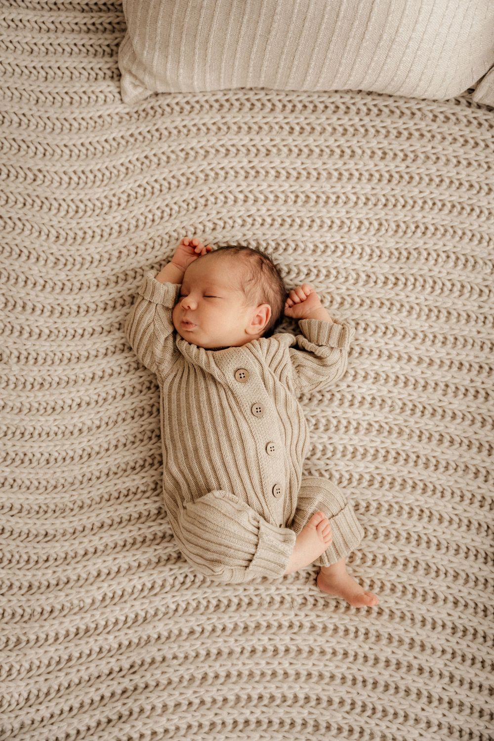 Newborn baby sleeping peacefully on a textured knit blanket wearing a beige ribbed button-up romper.