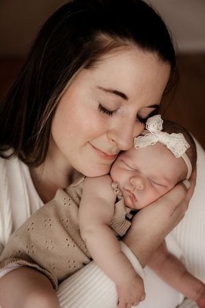 A smiling mother gently cradles her sleeping newborn baby girl wearing a lace bow headband and beige romper.