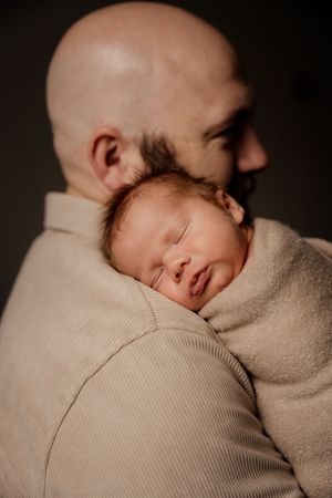 Bald father tenderly holding sleeping newborn baby wrapped in beige blanket against his shoulder.