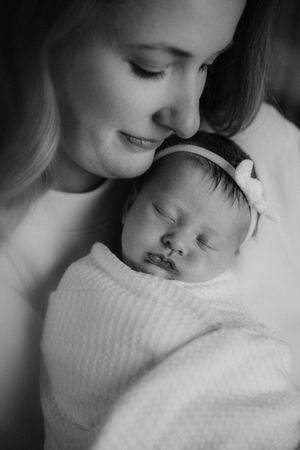 Black and white photo of a mother tenderly holding her sleeping swaddled newborn baby girl wearing a bow headband.