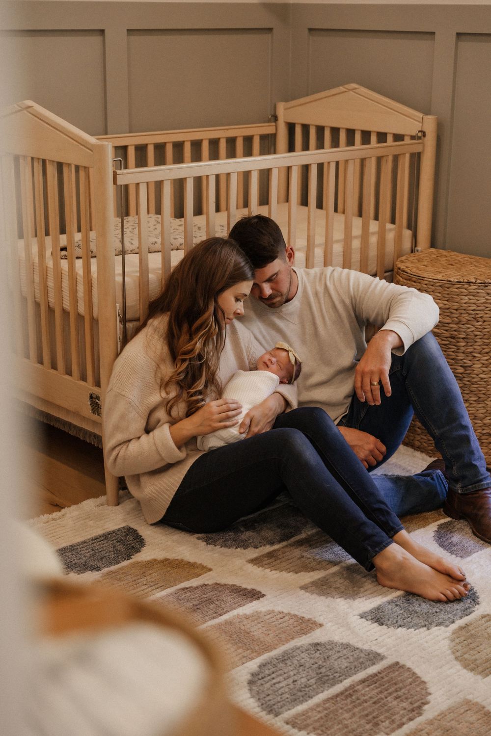 A couple sits on a nursery floor holding their newborn baby in front of a wooden crib.