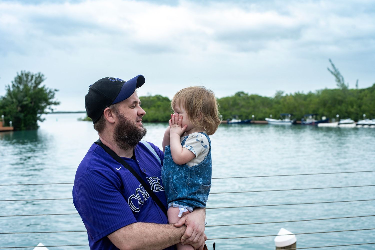 A bearded man in a blue cap holds a young child near a scenic waterway with lush green trees in the background.