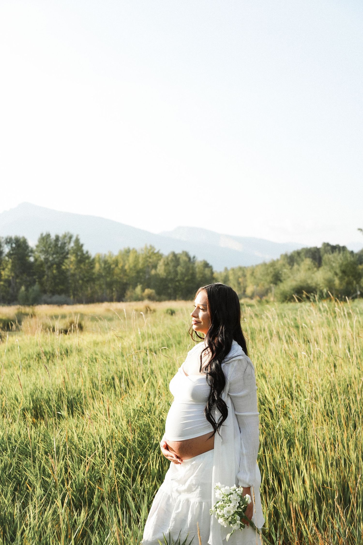Pregnant woman in white dress holding flowers in a green meadow with mountains and trees in the background.