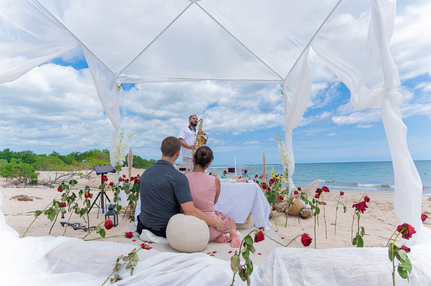 Man proposes to woman on one knee at a romantic beach sunset, holding her hand with golden skies and ocean waves behind them.