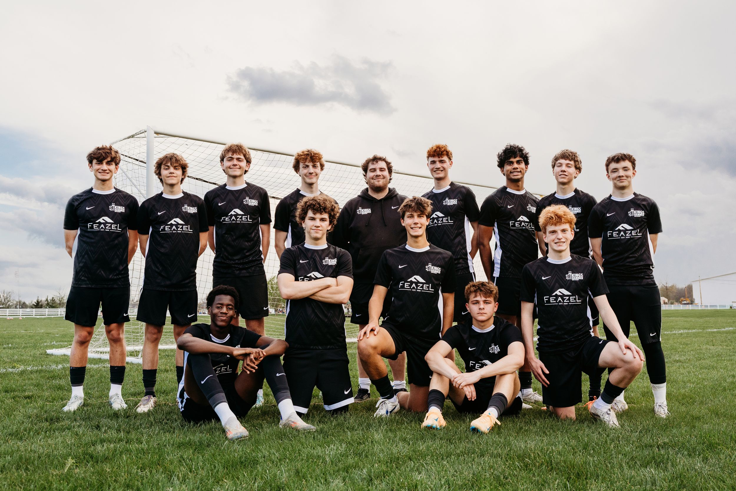 Youth soccer team poses in black uniforms on a green field with a goal net visible in the background.
