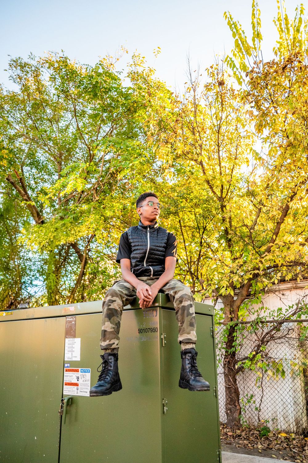 Young man in camo pants and black boots sitting atop a green utility box surrounded by autumn trees.
