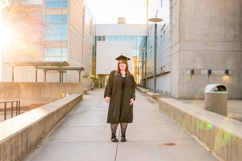 Graduate in cap and gown poses outdoors on campus walkway during golden hour sunset photography session.