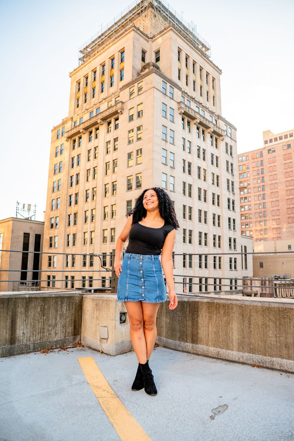 Young woman in denim skirt and black top posing on rooftop with classic urban building backdrop.