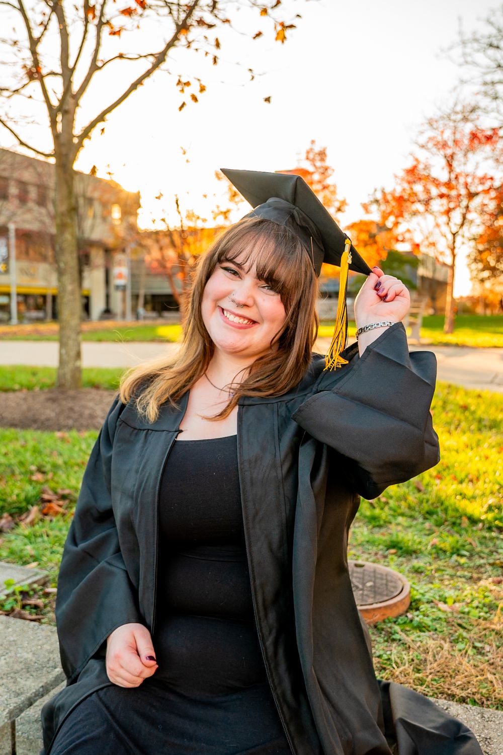 Smiling graduate in black cap and gown holding tassel outdoors during golden hour autumn setting.
