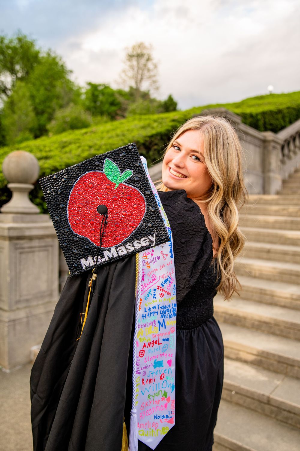 Smiling graduate in black gown displays decorated cap reading Ms. Massey with red apple design on outdoor steps.