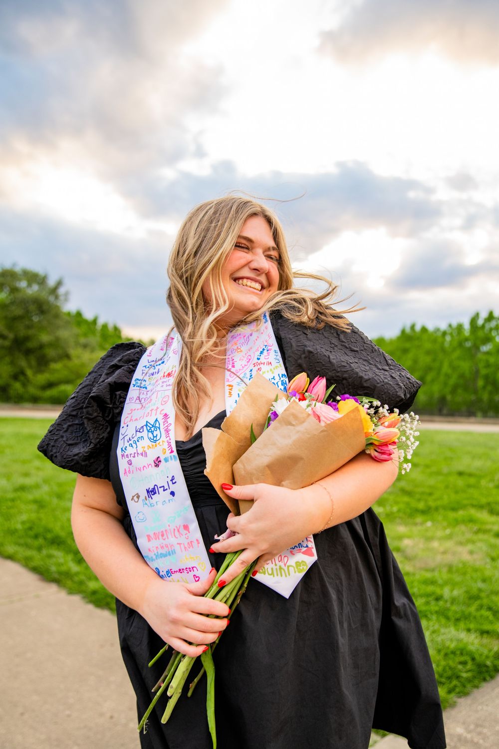 Smiling graduate in black dress holds flower bouquet, wearing colorful sash outdoors on cloudy day.