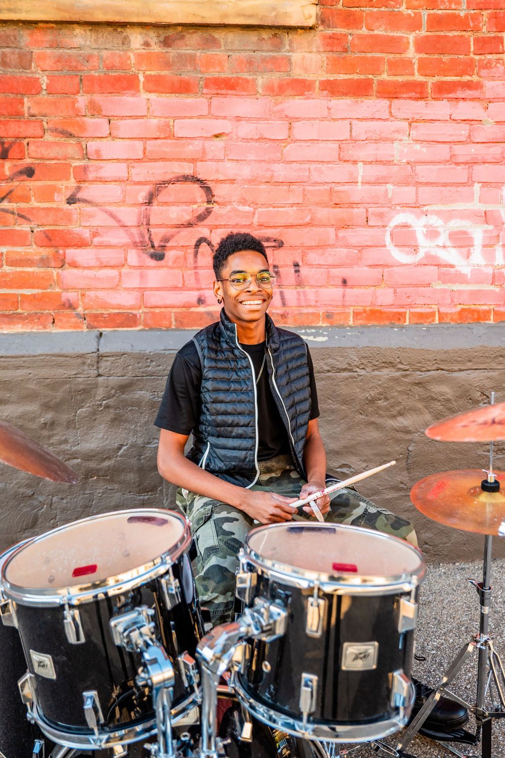 Smiling young drummer playing drums outdoors against a red brick wall with graffiti, wearing sunglasses and a vest.