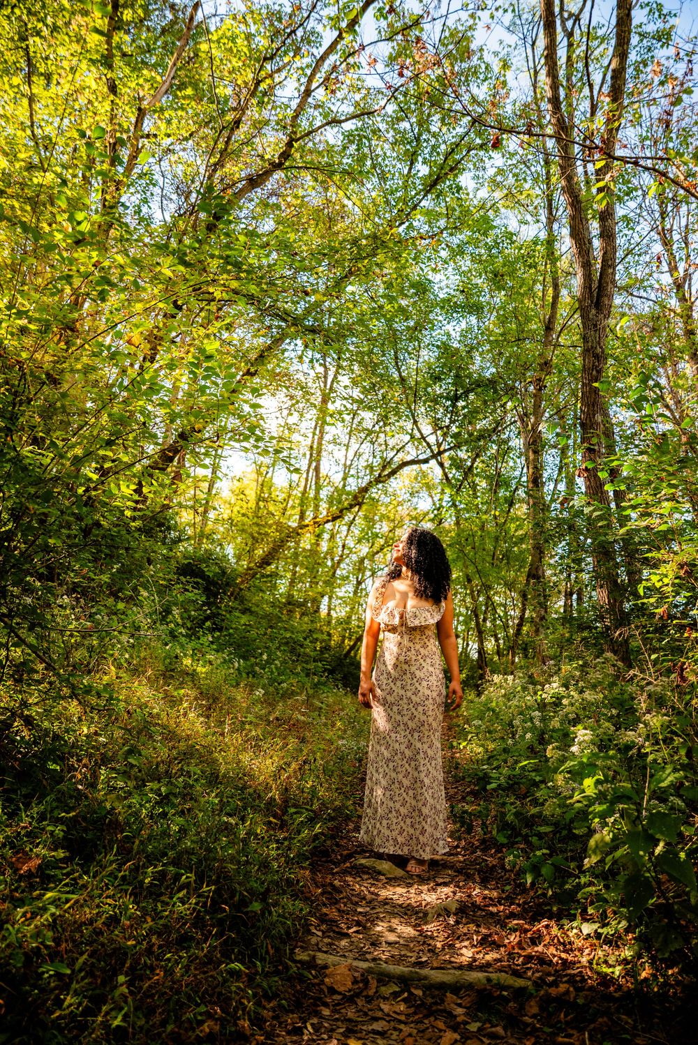 Woman in floral maxi dress walking along a sunlit forest path surrounded by lush green trees and foliage.