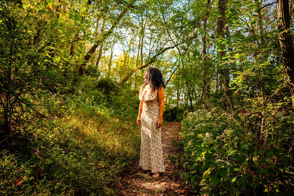 A woman in a floral dress stands on a sunlit forest path surrounded by lush green trees and foliage.