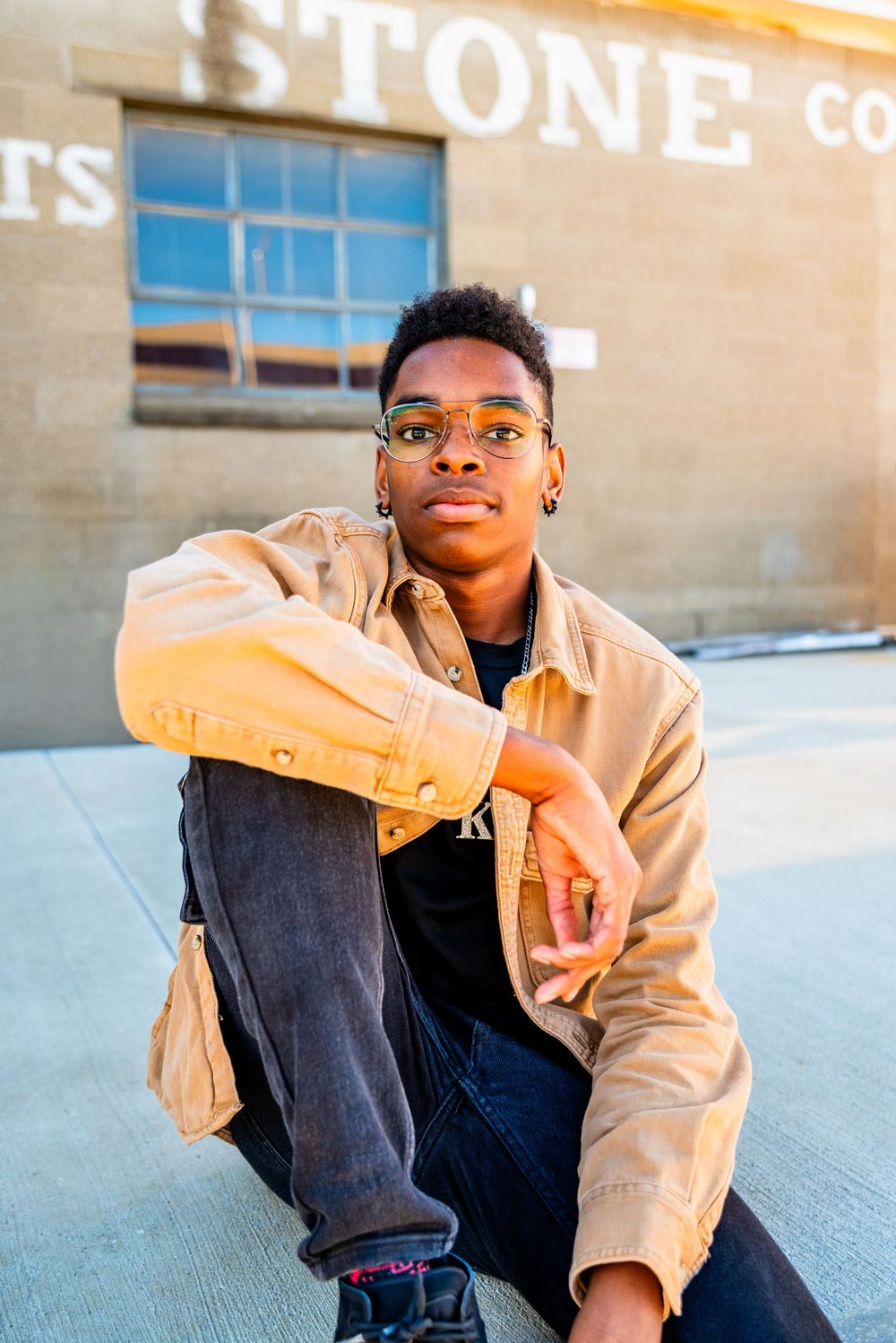 Young Black man in tan jacket and glasses sits confidently outdoors near a stone building in golden hour light.