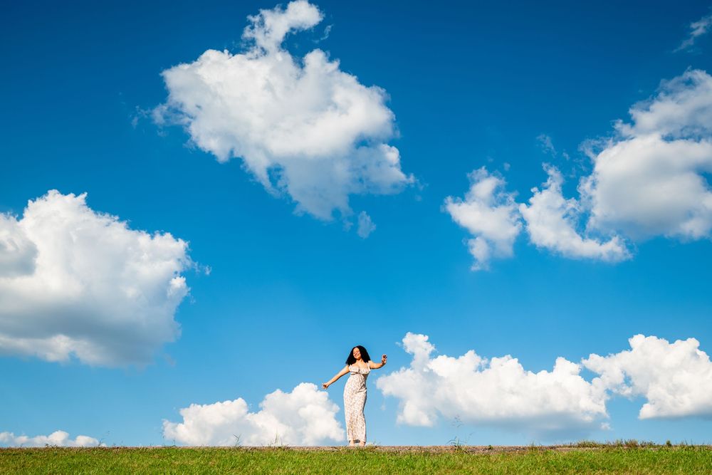 Woman in white dress standing on green hill under vibrant blue sky with fluffy white clouds on sunny day.