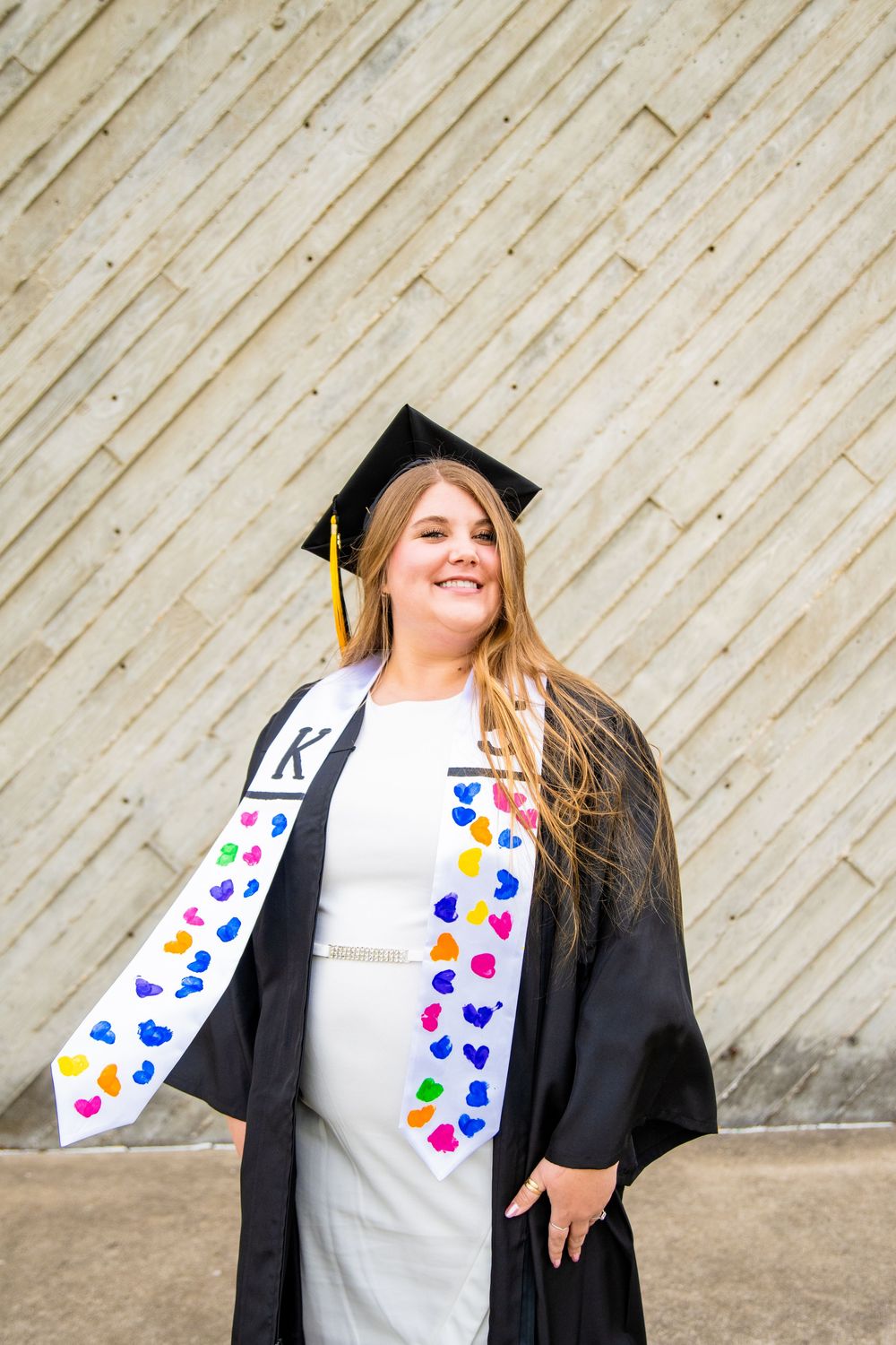 Smiling female graduate in black cap and gown with colorful heart-decorated stole posing outdoors.