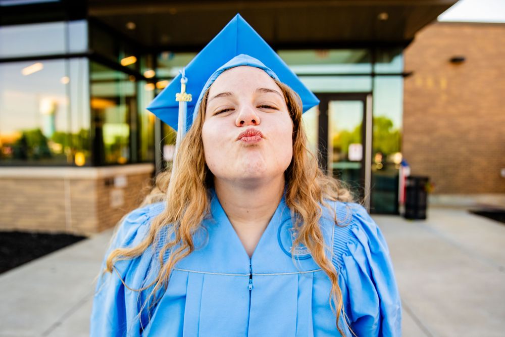 A young woman in blue graduation cap and gown making a kissy face outdoors near a brick building.