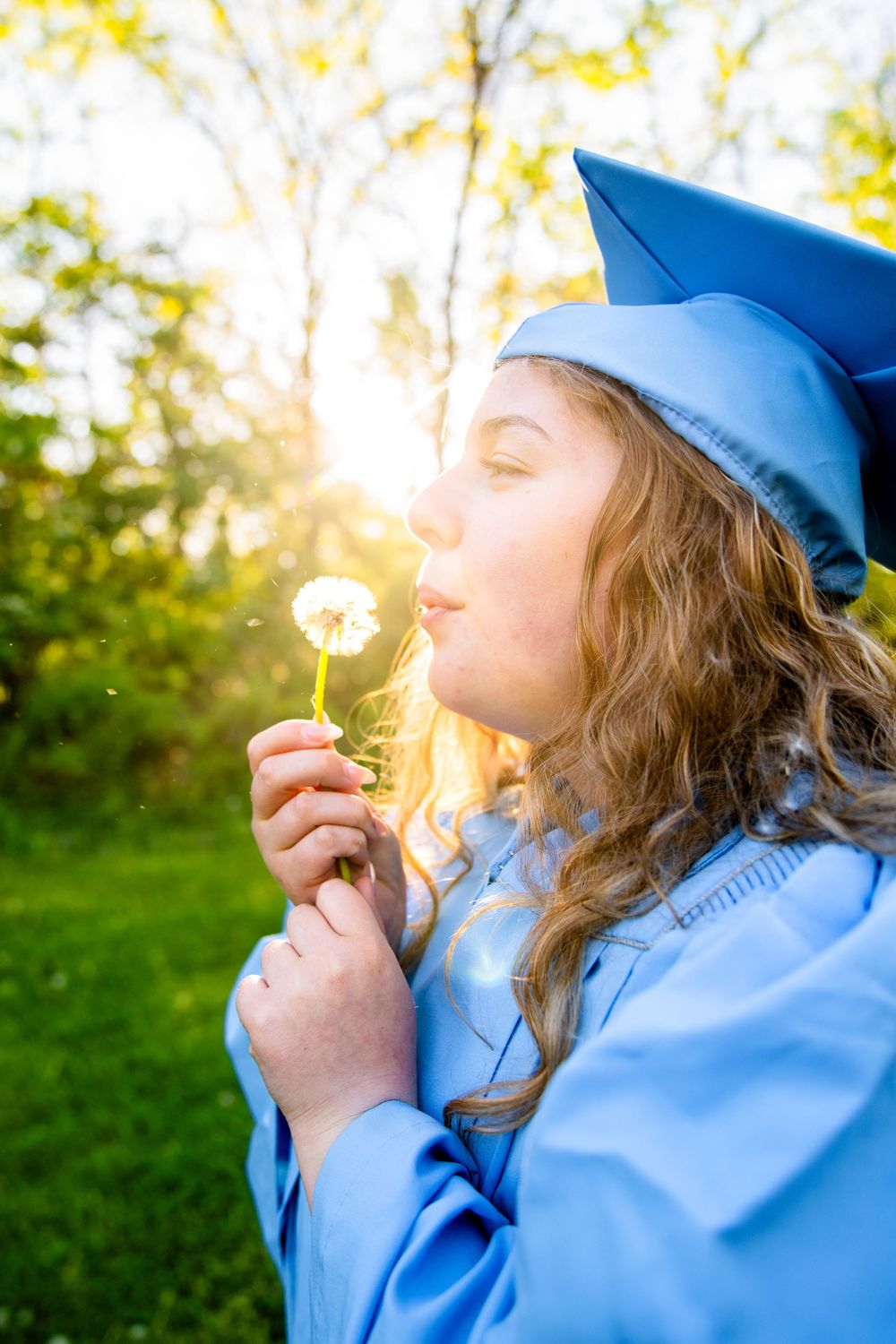 Girl in blue graduation cap and gown blowing dandelion seeds outdoors in golden sunlight.