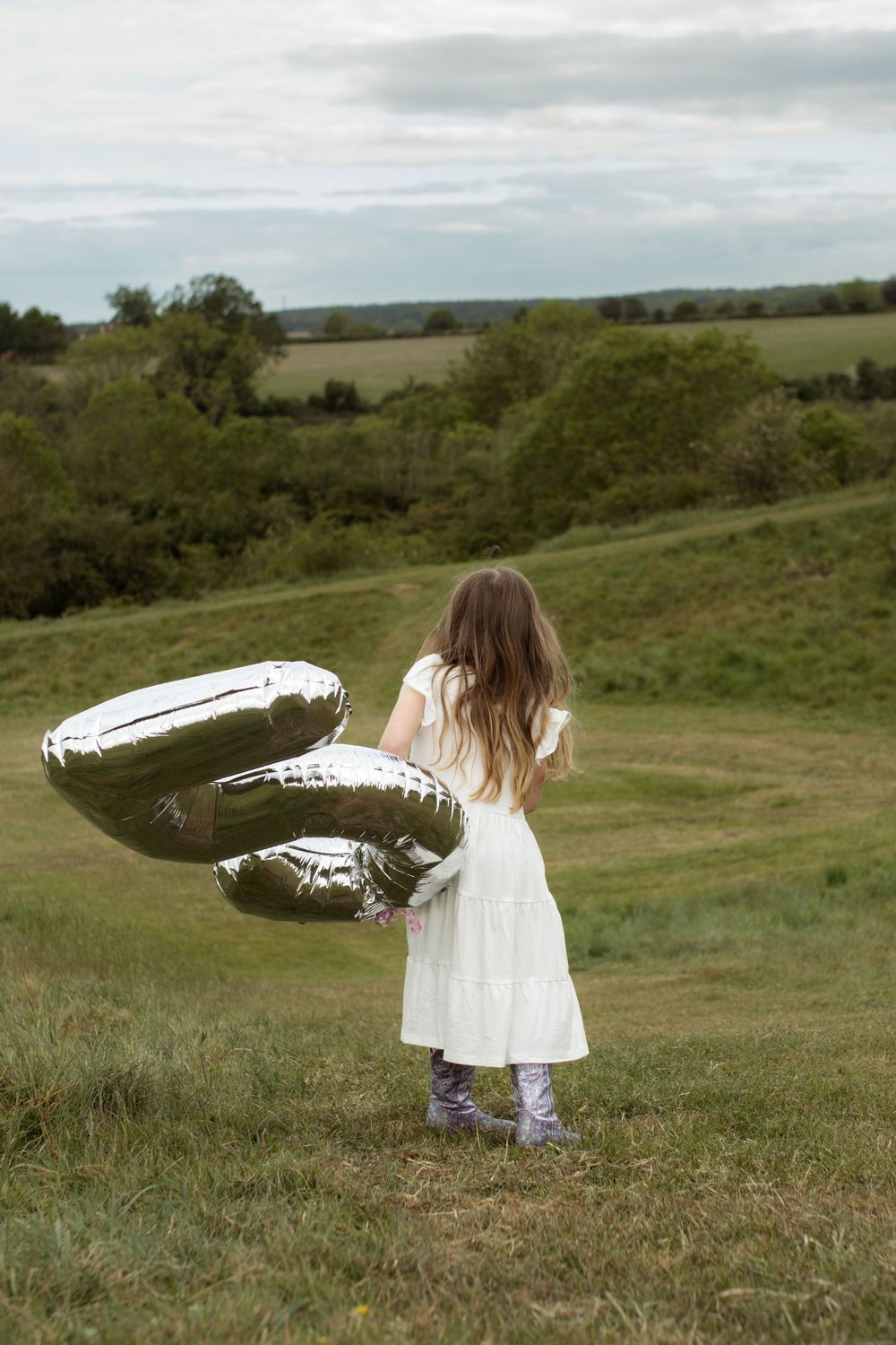 Young girl in white dress holding silver balloons stands in lush green field with trees in background.