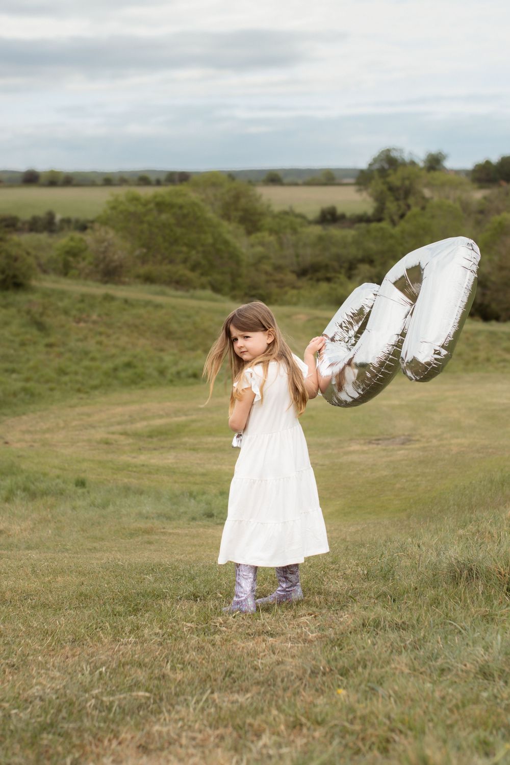 Young girl in white dress holding silver balloon in green countryside field.