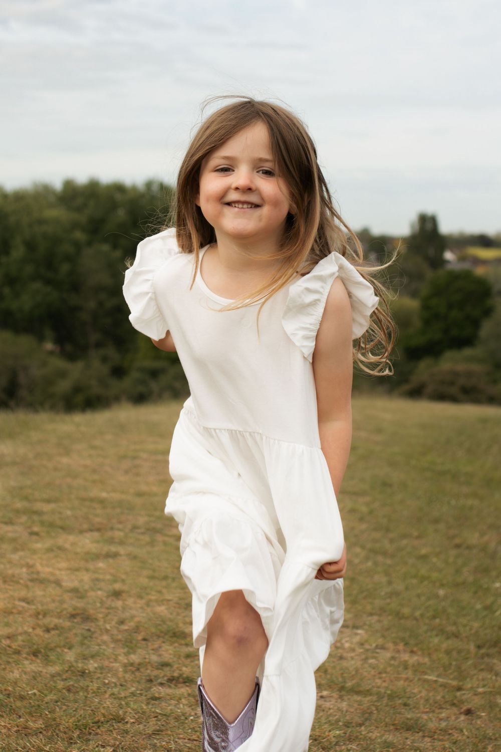 A joyful young girl in a white ruffled dress runs through a green field, smiling brightly on a cloudy day.