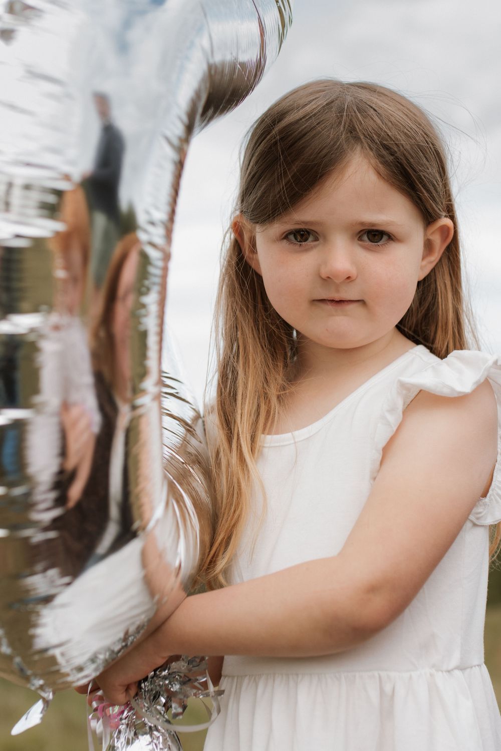 Young girl in white dress holding a large silver balloon, looking at camera with a serious expression.