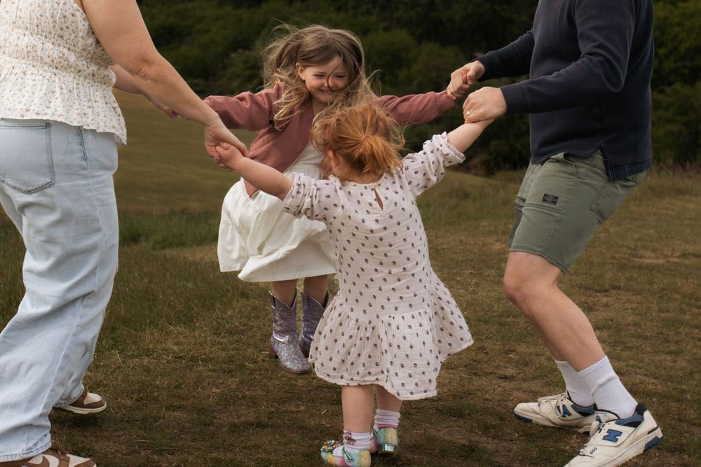 Two young girls hold hands with adults playing ring-around-the-rosy in a green field outdoors.