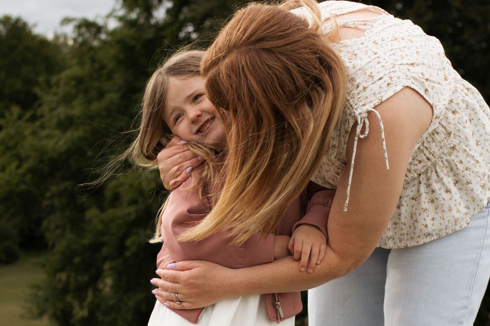 A mother with red hair hugs her smiling young daughter outdoors near green trees.