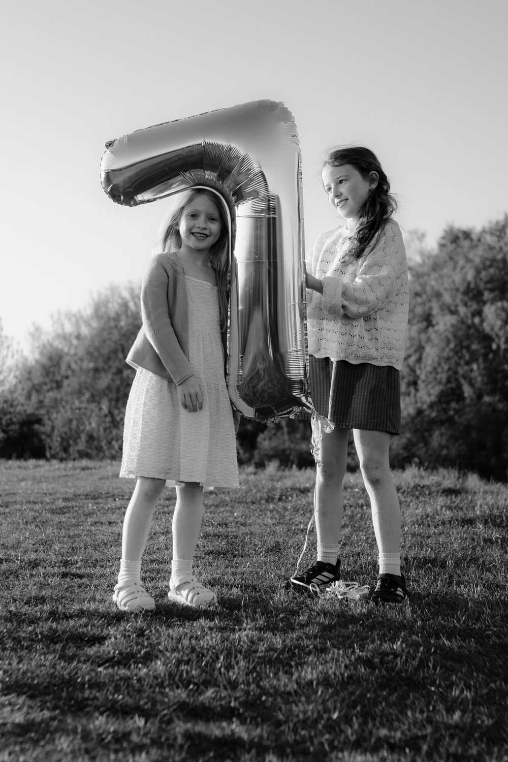 Two young girls smiling and holding a large number 7 balloon together on a sunny lawn.