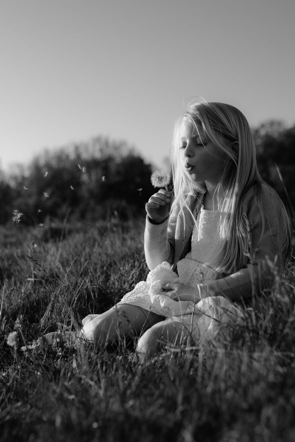 Black and white photo of a young girl sitting in grass, blowing dandelion seeds on a sunny day.