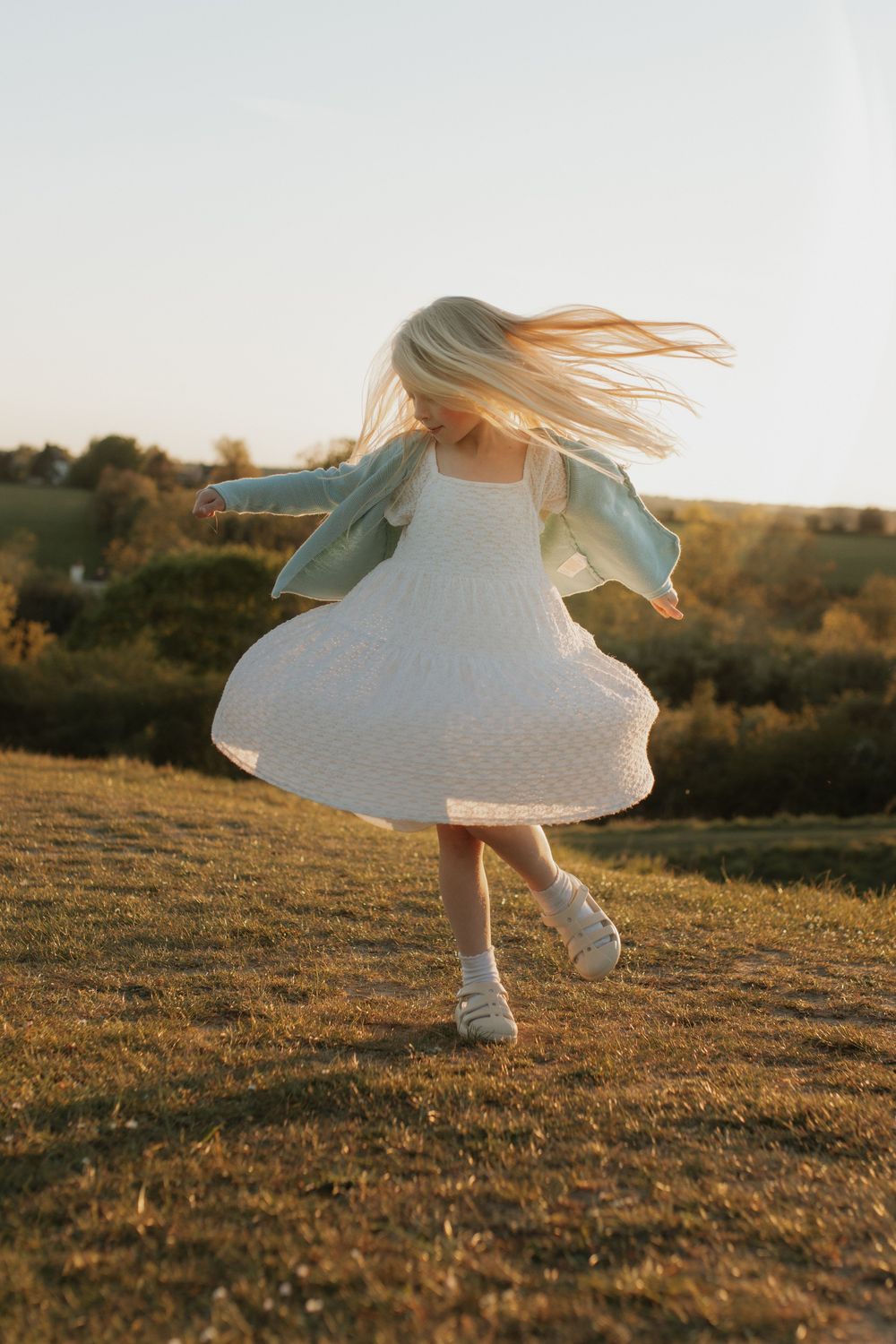 A young blonde girl twirls in a white dress and blue jacket on a sunlit grassy hill at golden hour.