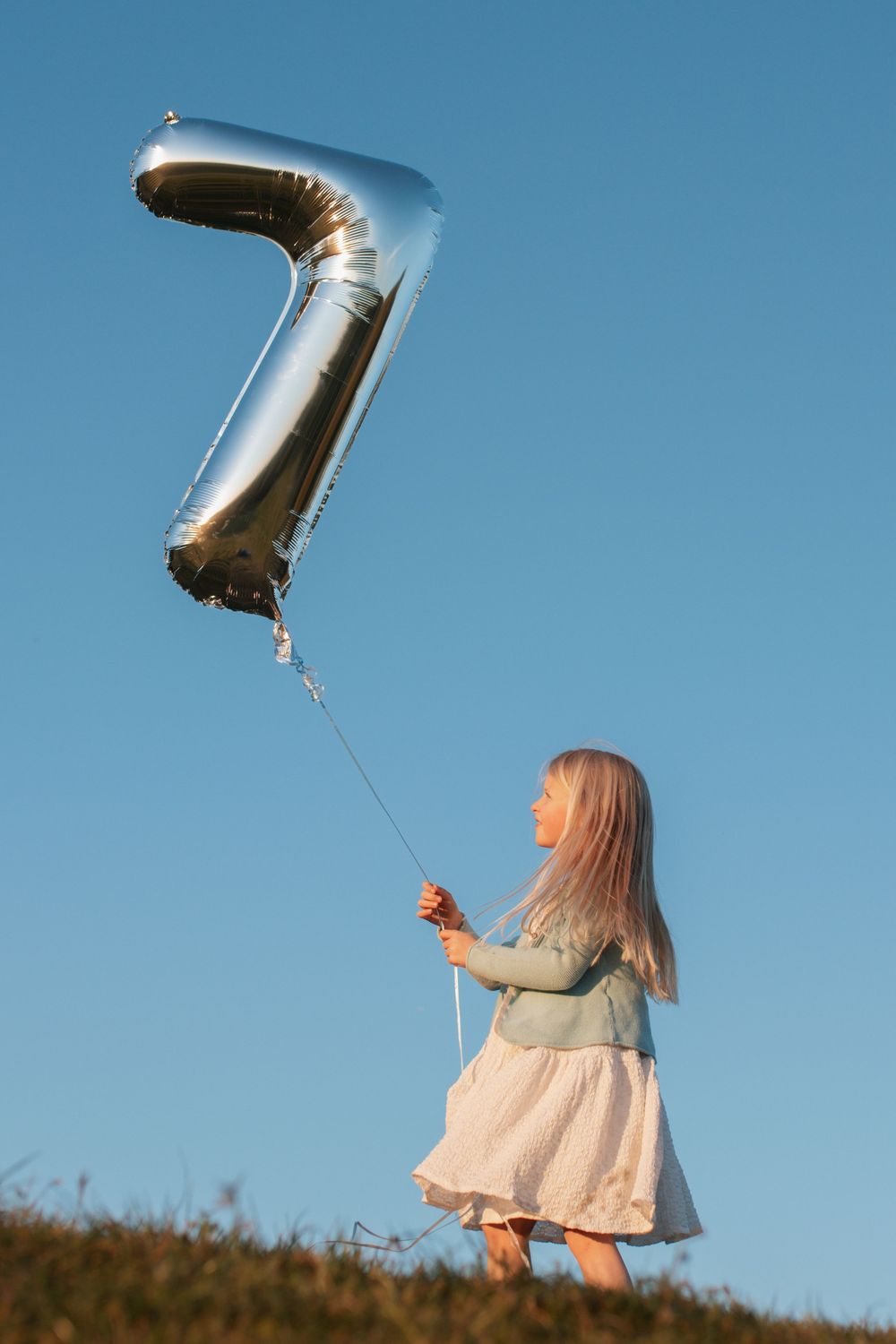 Young girl holding a silver number 7 balloon outdoors against a clear blue sky on a grassy hill.
