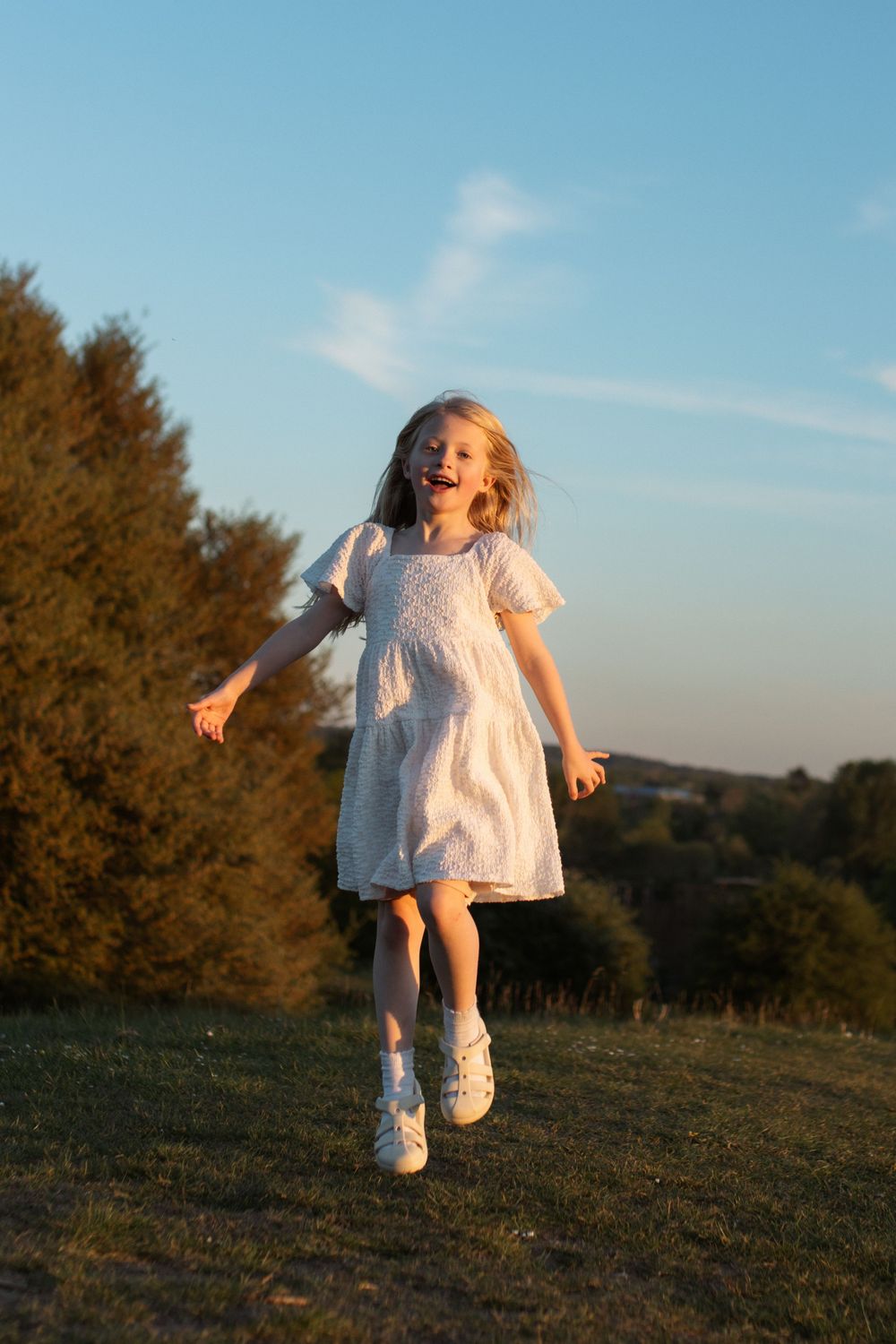 A joyful young girl in a white dress runs through a sunny green field at golden hour with trees in the background.