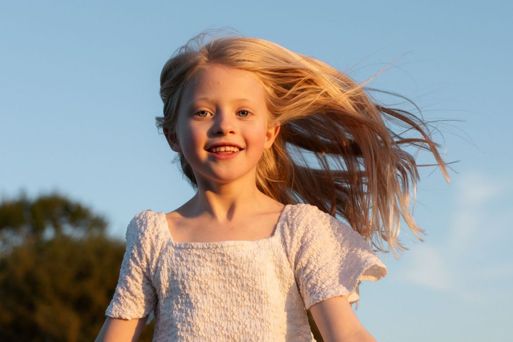 Happy young blonde girl with hair blowing in the wind, smiling outdoors against a blue sky.
