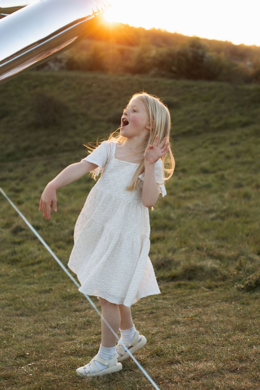 A joyful blonde girl in a white dress plays outdoors at golden sunset on a green hillside.